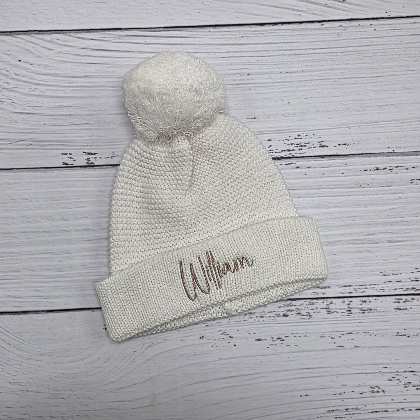 Knitted beanie with pom-pom and embroidered name on a wooden surface