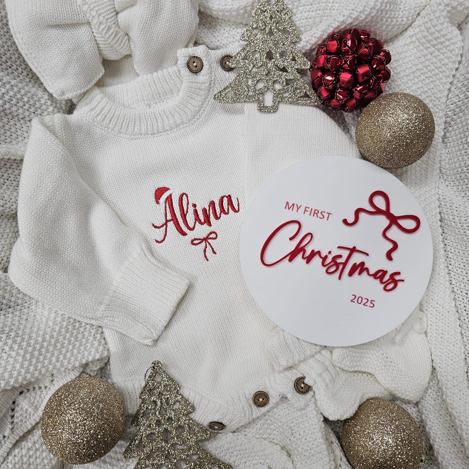 Baby sweater with 'Alina My First Christmas 2025' text, surrounded by Christmas decorations on a textured surface.