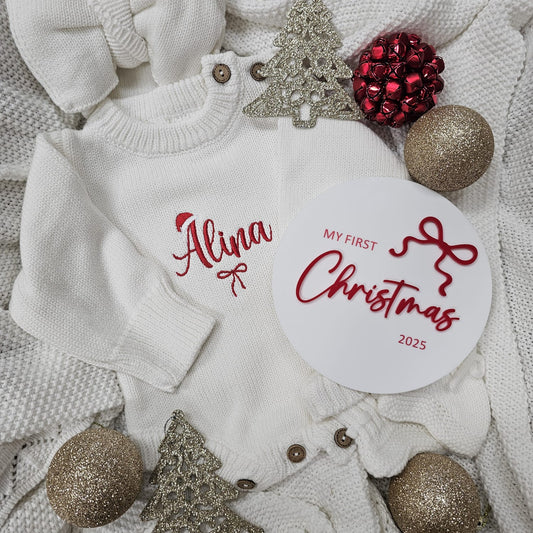 Baby sweater with 'Alina My First Christmas 2025' text, surrounded by Christmas decorations on a textured surface.