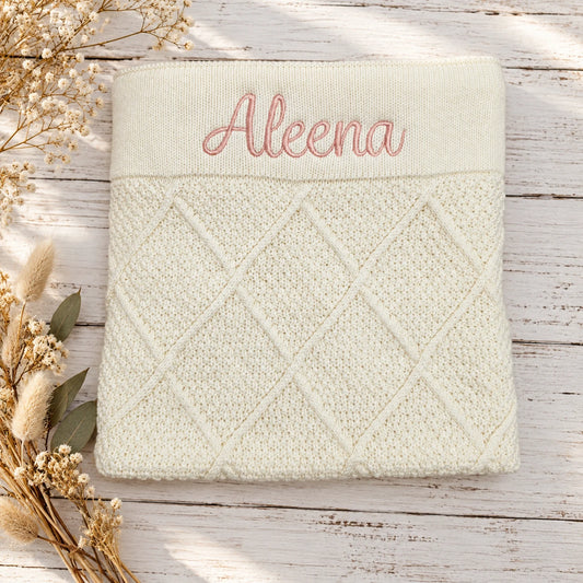 A folded cream-colored knit blanket with a diamond cable pattern and personalized pink embroidery reading 'Aleena' on the top band, displayed on a rustic white wooden surface with dried flowers.