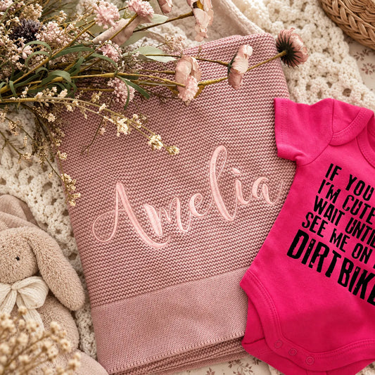 A styled flat-lay of a pink knit blanket with the name 'Amelia' embroidered in light pink script, arranged on a cream crochet blanket with dried flowers, a beige bunny toy, and a bright pink baby bodysuit with black text.