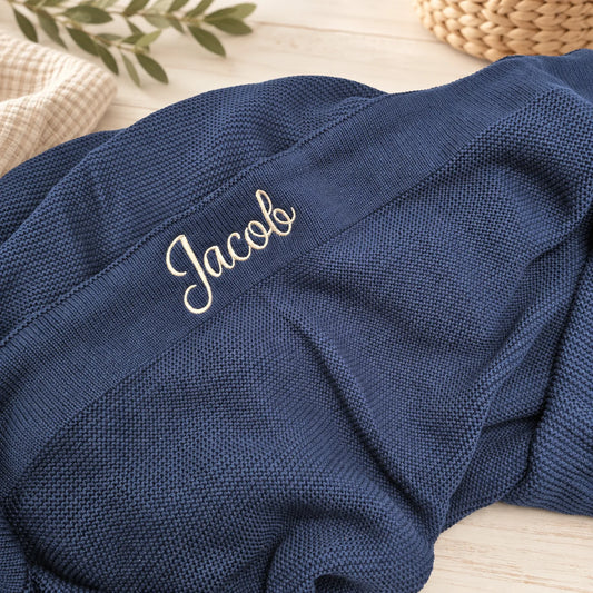 A close-up of a navy blue textured knit blanket with the name 'Jacob' embroidered in white script, styled on a light wood surface with decorative props.