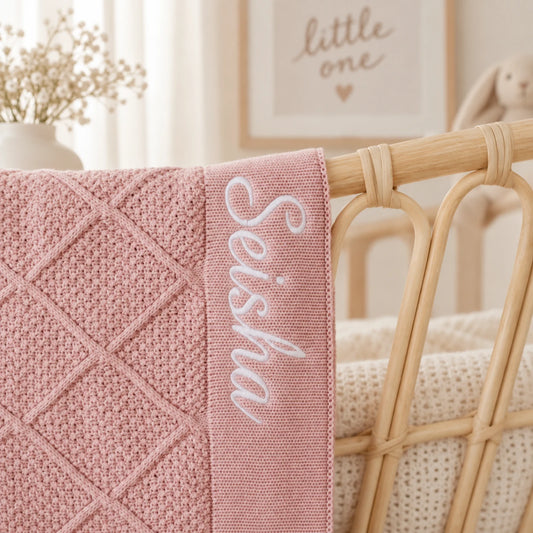A close-up of a dusty pink knitted baby blanket with a diamond quilted pattern and white embroidered script reading 'Seisha', draped over a light wood rattan crib in a softly lit nursery.