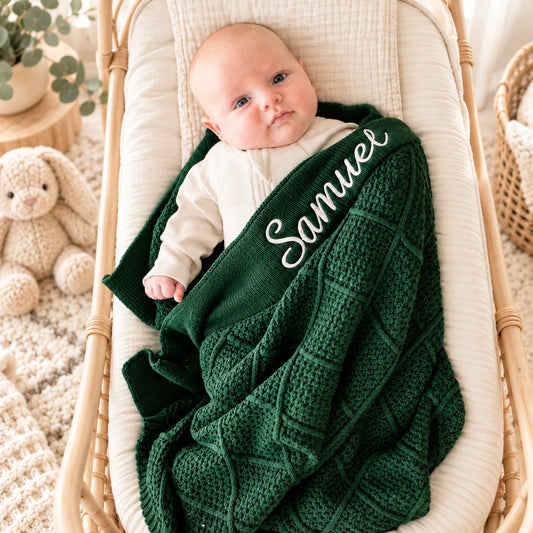 A baby wrapped in a dark green cable-knit blanket with the name 'Samuel' embroidered in white script, resting in a woven bassinet.