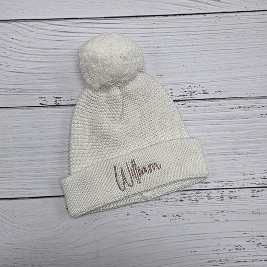 Knitted beanie with pom-pom and embroidered name on a wooden surface