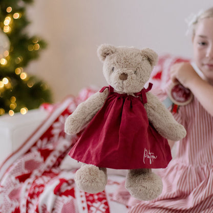 Child holding a teddy bear in a red dress with a Christmas tree in the background