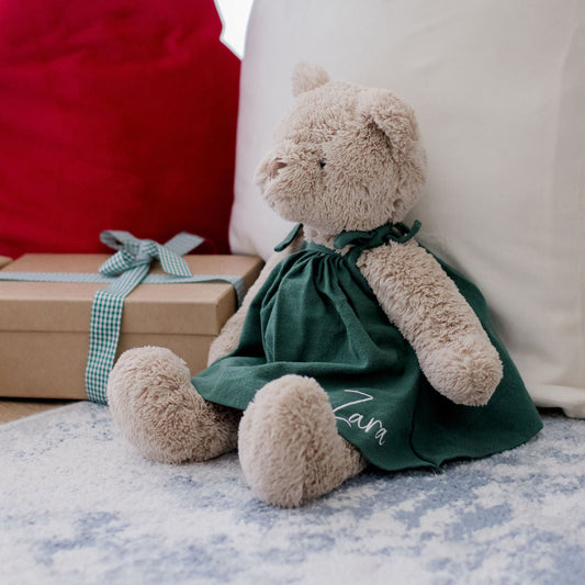 Teddy bear in a green dress sitting next to a gift box with a red bag and white drawstring bag in the background.