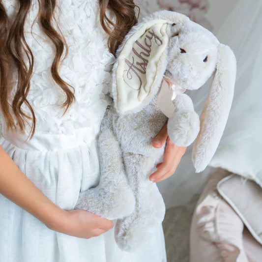 Child holding a plush bunny toy with 'Holly' embroidered on it, wearing a white dress.