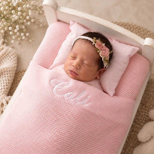 A newborn baby is sleeping peacefully in a white wooden bassinet, wrapped in a pink knit blanket with the name 'Cali' embroidered in white script. The baby wears a pink floral headband, resting on a pink pillow.