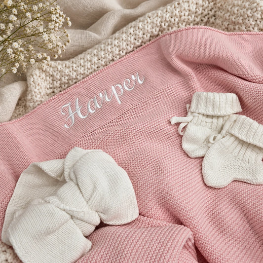 A close-up flat lay of a pink knit blanket with the name 'Harper' embroidered in white script across the waistband. White knit baby booties and a matching cream knit blanket are arranged on top, with small white flowers in the upper left corner.