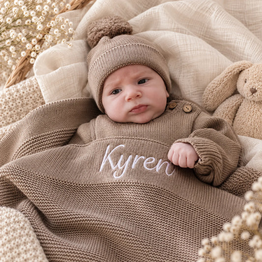 A baby lying on a soft cream blanket, wearing a knitted pom-pom beanie and a matching long-sleeve knit outfit with the name 'Kyren' embroidered in white across the chest. A beige teddy bear and dried floral decor are visible in the scene.