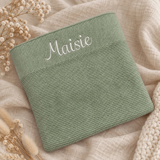 A sage green knit cotton blanket with the name 'Maisie' embroidered in white script across the top, styled on a beige linen fabric with dried flowers.