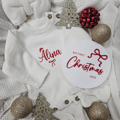 Baby sweater with 'Alina My First Christmas 2025' text, surrounded by Christmas decorations on a textured surface.
