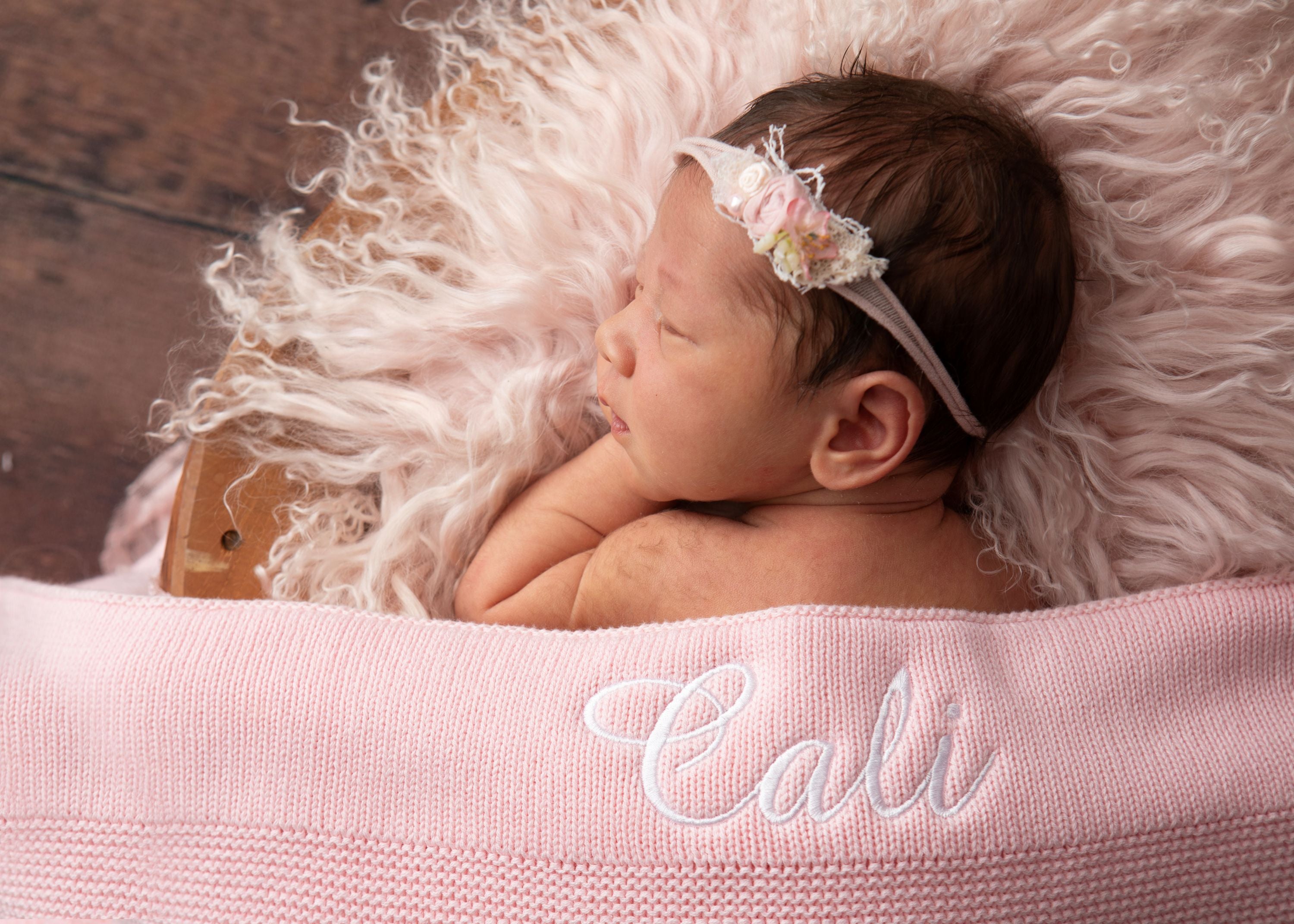 Newborn baby lying on a pink blanket with 'Cali' embroidered, wearing a floral headband.