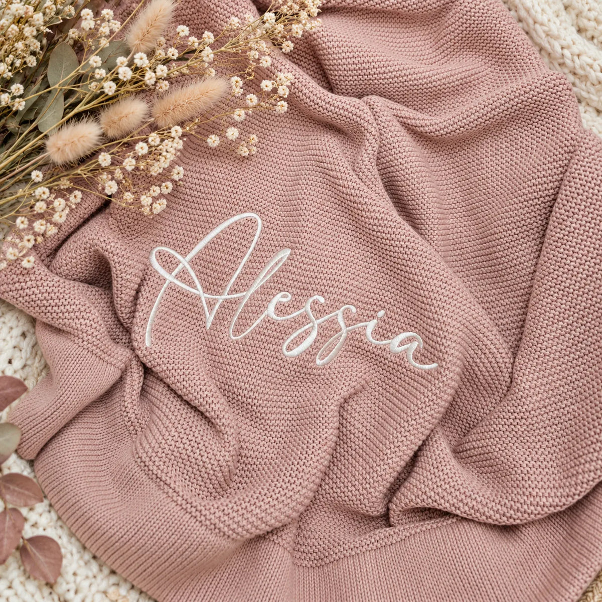 A dusty rose pink knit blanket with the name 'Alessia' embroidered in white script, styled with dried flowers on a white knit surface.