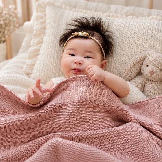 A baby lying on a cream knit blanket with a plush bunny, covered by a dusty pink knitted blanket personalized with the name 'Amelia' in white script. The baby wears a gold and pearl headband.