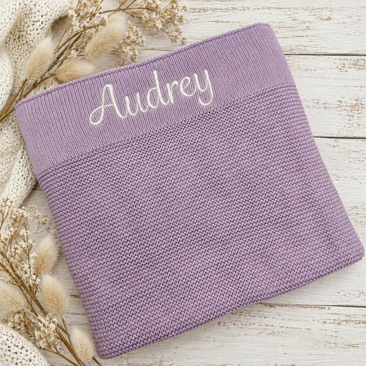 A folded knitted purple blanket with the name 'Audrey' embroidered in white script on the top edge, styled on a white wooden surface with dried flowers and a cream knit sweater.