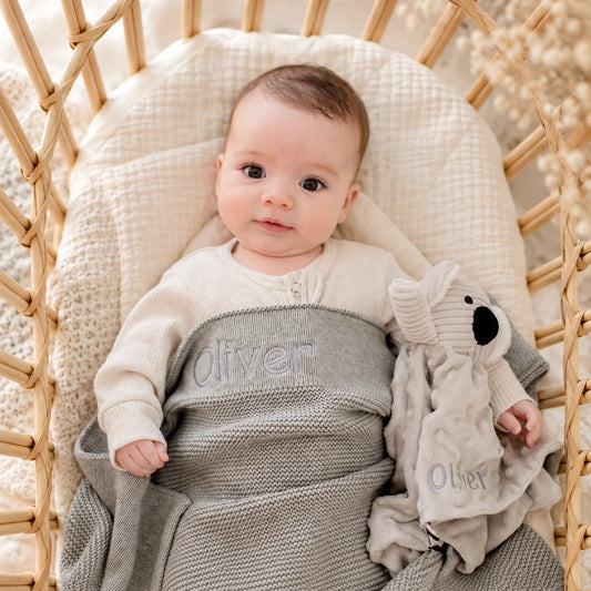 A baby lying in a woven rattan bassinet, wrapped in a gray knit blanket with the name 'Oliver' embroidered across the chest, holding a matching koala-shaped comforter. The baby is wearing a cream long-sleeve top.