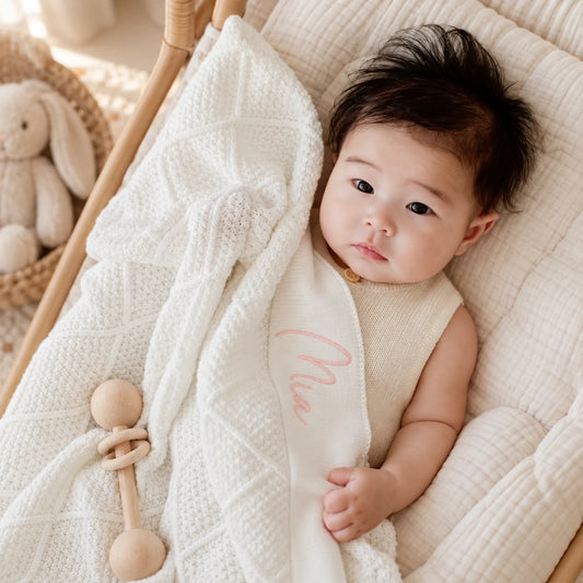 A baby lying on a cream-colored textured blanket inside a wooden bassinet, wearing a cream sleeveless top with a pink embroidered script.