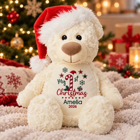 A plush cream teddy bear wearing a red Santa hat sits on a soft blanket in front of a blurred Christmas tree and gift box. The bear has a custom cream bib printed with 'My 1st Christmas Amelia 2026' in red and green.