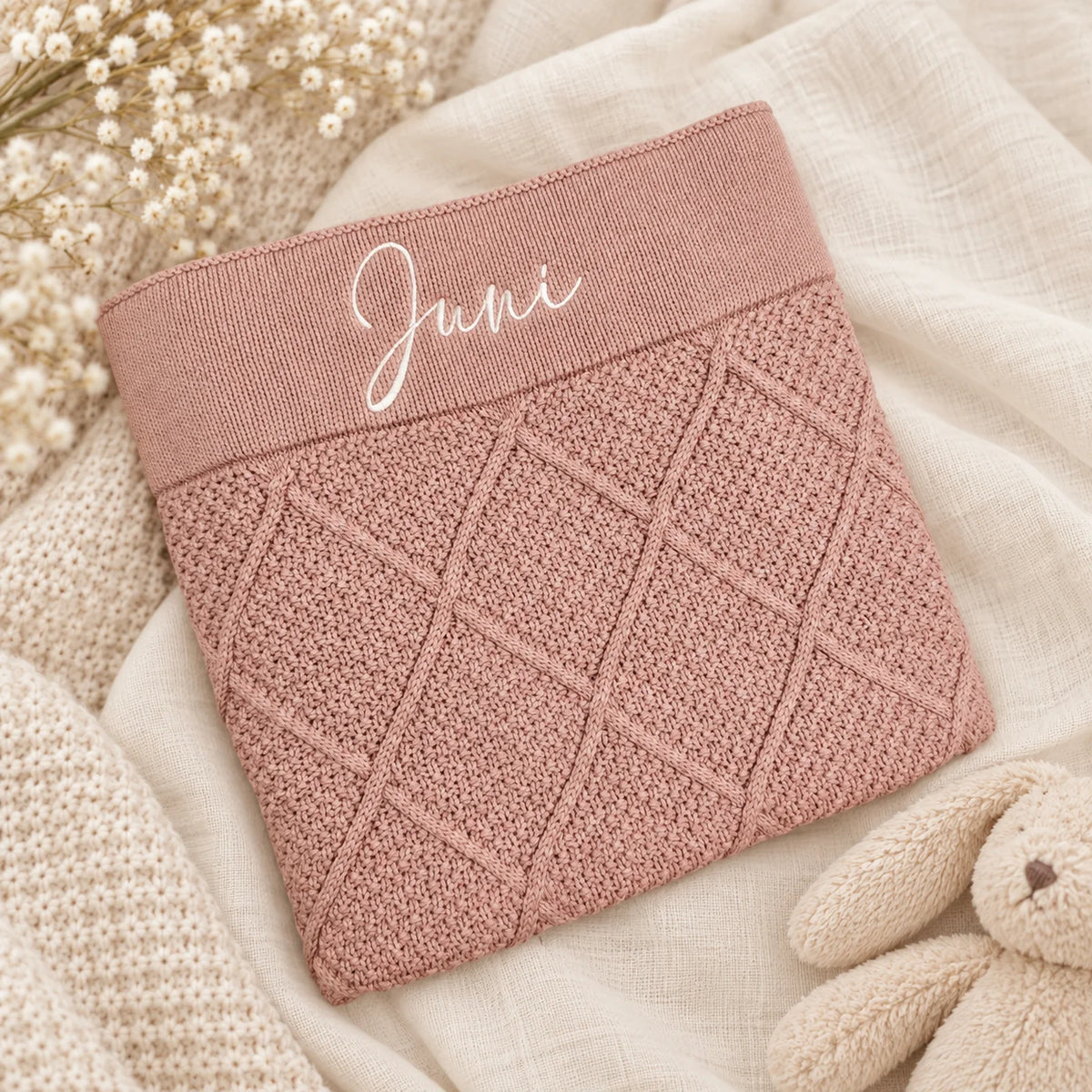 A dusty pink knit baby blanket with a diamond cable pattern and a ribbed top band embroidered with the name 'Juni' in white script, styled on a cream linen fabric with dried flowers and a plush bunny.