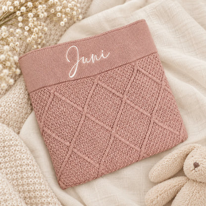 A dusty pink knit baby blanket with a diamond cable pattern and a ribbed top band embroidered with the name 'Juni' in white script, styled on a cream linen fabric with dried flowers and a plush bunny.