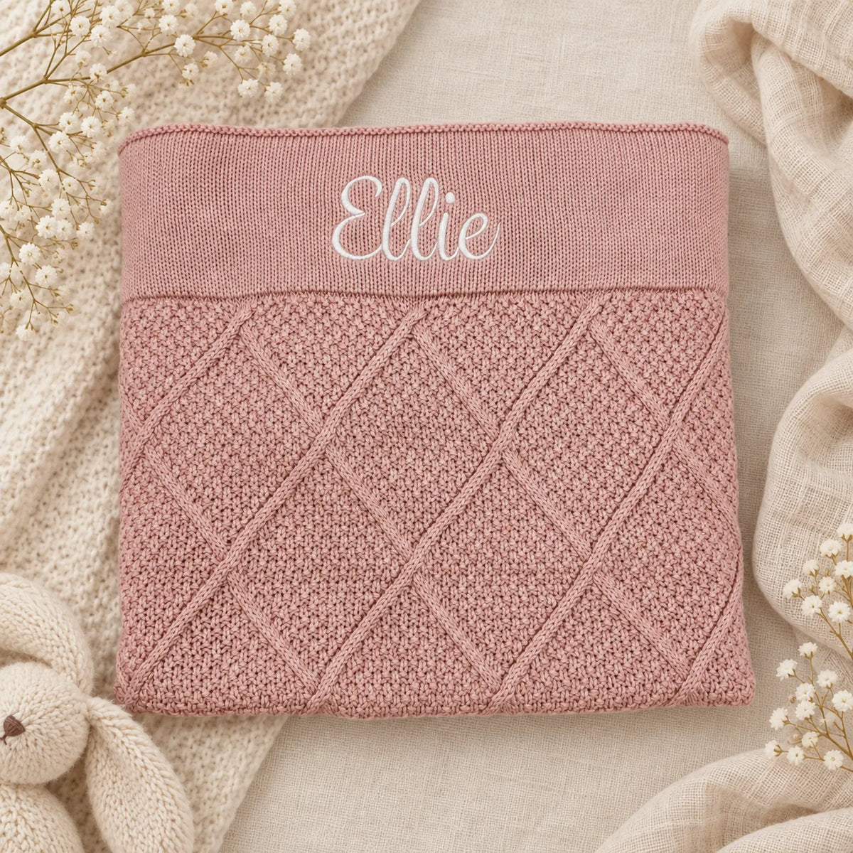 A folded dusty pink knit blanket with a diamond cable-knit pattern and the name 'Ellie' embroidered in white script on the top band, styled on a neutral beige fabric background with dried flowers and a knit throw.