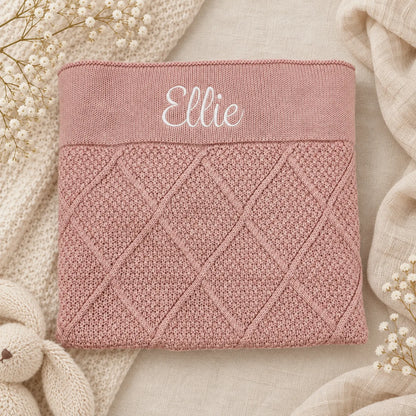A folded dusty pink knit blanket with a diamond cable-knit pattern and the name 'Ellie' embroidered in white script on the top band, styled on a neutral beige fabric background with dried flowers and a knit throw.