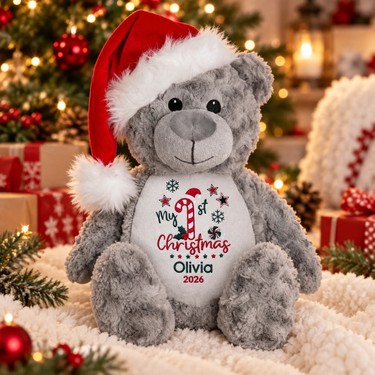 A gray plush teddy bear wearing a red Santa hat sits on a white textured blanket in a festive holiday setting with Christmas trees, ornaments, and wrapped gifts in the background. The bear's belly is printed with 'My 1st Christmas Olivia 2026' in red and green text with candy cane and snowflake graphics.