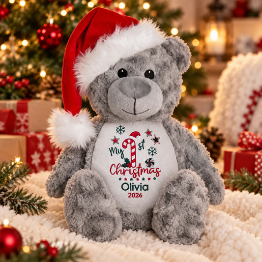 A gray plush teddy bear wearing a red Santa hat sits on a white textured blanket in a festive holiday setting with Christmas trees, ornaments, and wrapped gifts in the background. The bear's belly is printed with 'My 1st Christmas Olivia 2026' in red and green text with candy cane and snowflake graphics.