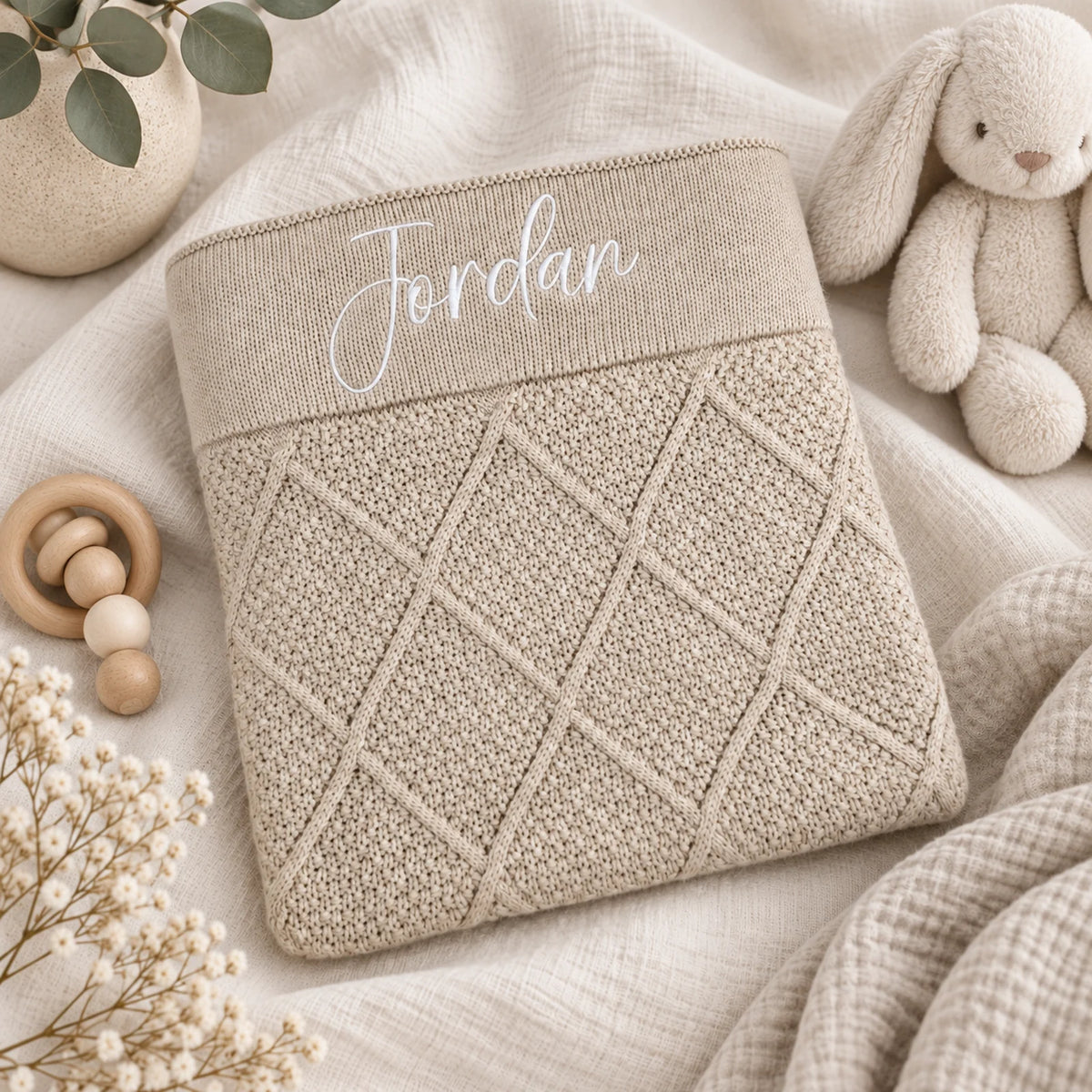 A beige knitted baby blanket or pouch with a diamond cable pattern and the name 'Jordan' embroidered in white script on the top cuff, styled on a soft cream fabric with a plush bunny, wooden teether, and dried flowers.