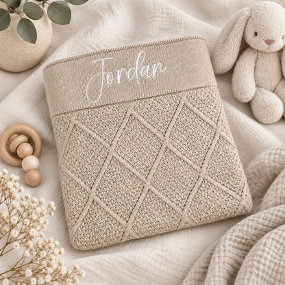 A beige knitted baby blanket or pouch with a diamond cable pattern and the name 'Jordan' embroidered in white script on the top cuff, styled on a soft cream fabric with a plush bunny, wooden teether, and dried flowers.