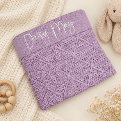 A lavender purple knitted baby blanket with a diamond cable pattern and a ribbed top band featuring the personalized name 'Daisy May' in white script, styled on a cream knit blanket with a plush bunny toy and wooden beads.