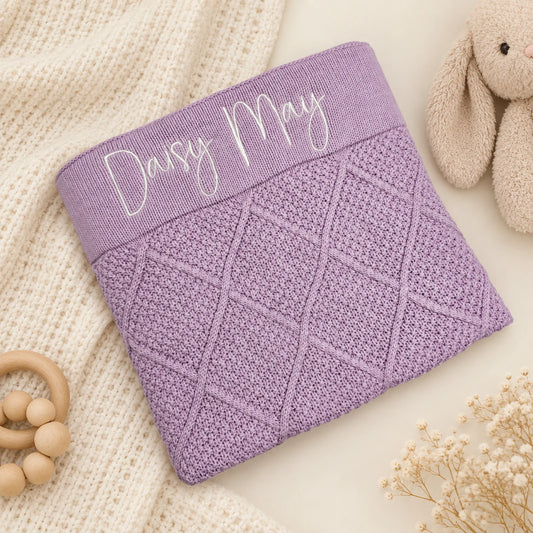A lavender purple knitted baby blanket with a diamond cable pattern and a ribbed top band featuring the personalized name 'Daisy May' in white script, styled on a cream knit blanket with a plush bunny toy and wooden beads.