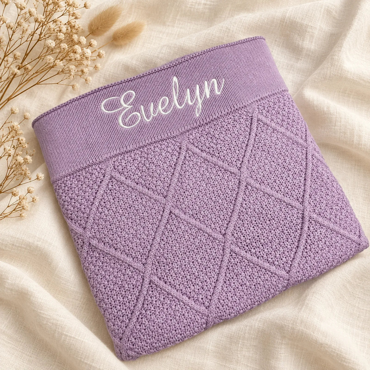 A folded lavender knit garment with a diamond cable-knit pattern and white embroidered name 'Evelyn' on the waistband, styled on a cream fabric background with dried flowers.