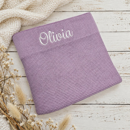 A lavender purple knit baby blanket with the name 'Olivia' embroidered in white script on the ribbed cuff, styled on a white wooden surface with a cream knit sweater and dried grasses.