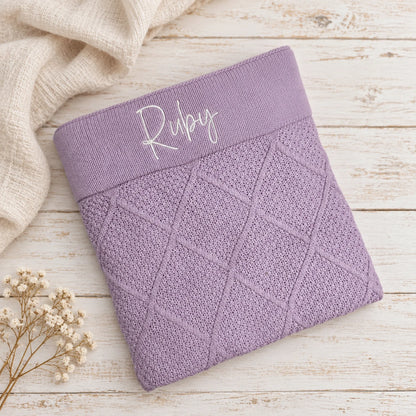 A folded lavender knit baby blanket or sweater with a diamond cable-knit pattern and a wide elastic band embroidered with the name 'Liana' in white script, shown on a beige textured background with dried grass stems.