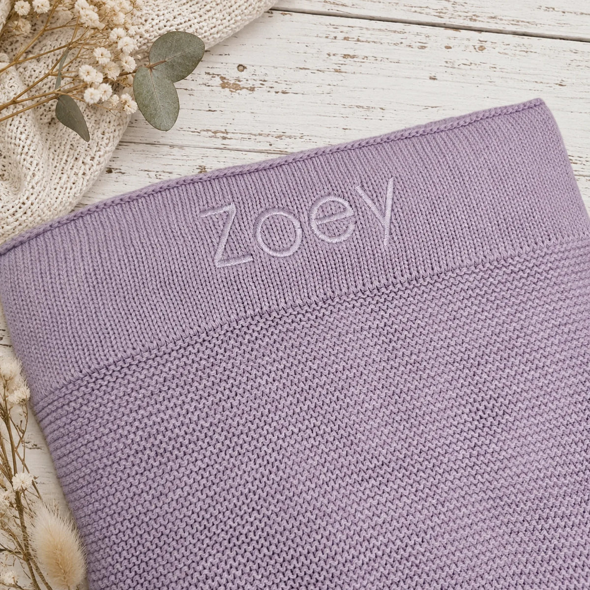 A close-up of a lavender knit blanket, with the name 'ZoeY' embroidered in lilac across the ribbed band. The item is styled on a white distressed wood surface with dried flowers and a cream knit fabric prop.