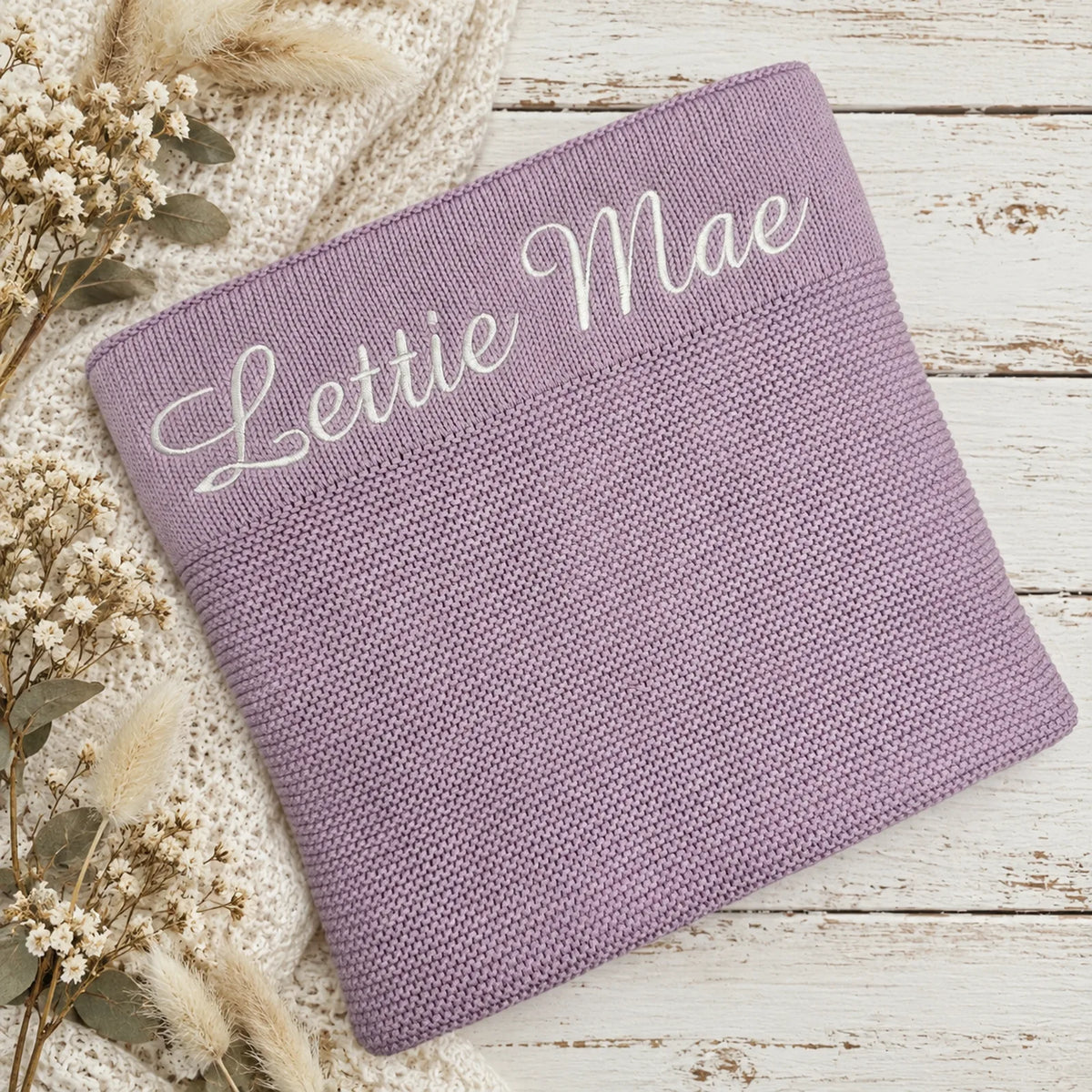 A personalized lavender knit blanket with 'Lettie Mae' embroidered in white script across the top, styled on a white wooden surface with a cream knit sweater and dried flowers.