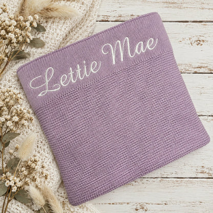 A personalized lavender knit blanket with 'Lettie Mae' embroidered in white script across the top, styled on a white wooden surface with a cream knit sweater and dried flowers.