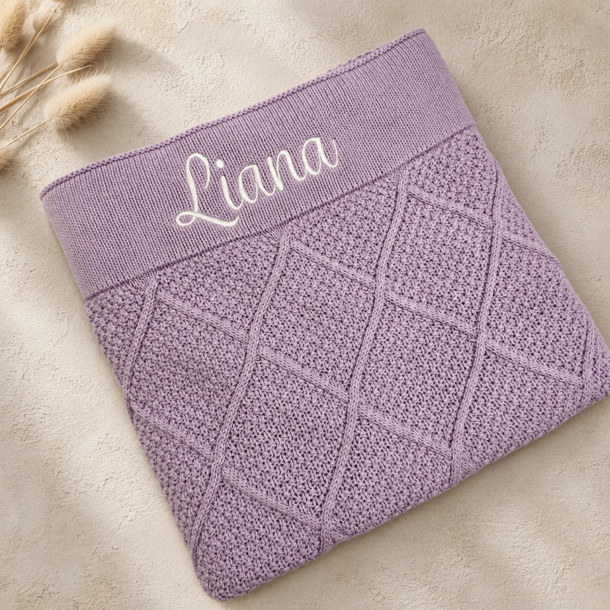 A folded lavender knit baby blanket or sweater with a diamond cable-knit pattern and a wide elastic band embroidered with the name 'Liana' in white script, shown on a beige textured background with dried grass stems.