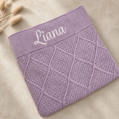 A folded lavender knit baby blanket or sweater with a diamond cable-knit pattern and a wide elastic band embroidered with the name 'Liana' in white script, shown on a beige textured background with dried grass stems.