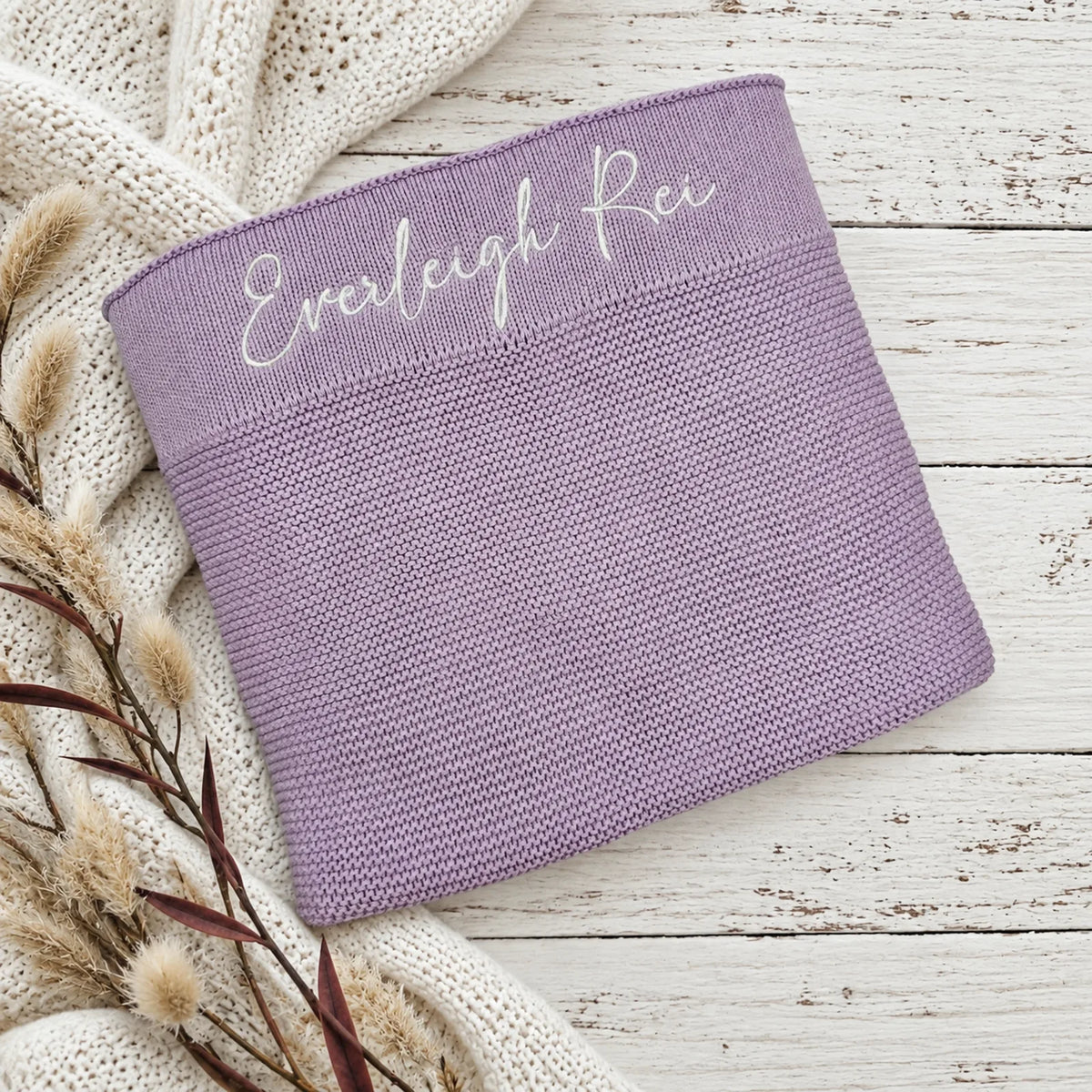 A lavender knit blanket with the name 'Everleigh Rei' embroidered in white script across the top, styled on a white wood surface with a cream knit blanket and dried grass stems.