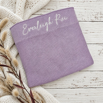 A lavender knit blanket with the name 'Everleigh Rei' embroidered in white script across the top, styled on a white wood surface with a cream knit blanket and dried grass stems.