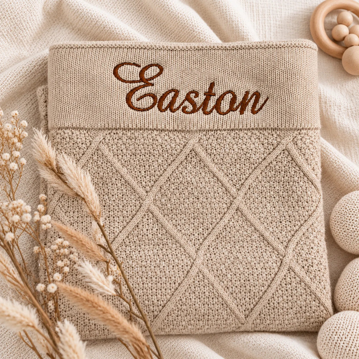 A folded beige knit baby blanket or swaddle with a diamond cable-knit pattern and the name 'Easton' embroidered in brown script across the top, styled on a soft cream fabric with dried grasses and wooden baby props.