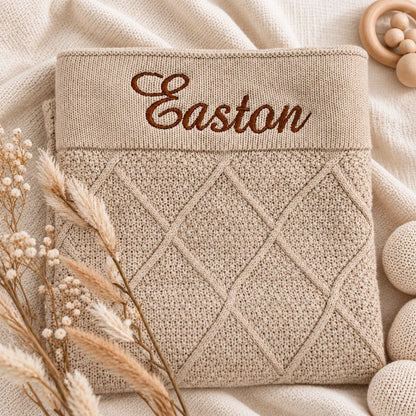 A folded beige knit baby blanket or swaddle with a diamond cable-knit pattern and the name 'Easton' embroidered in brown script across the top, styled on a soft cream fabric with dried grasses and wooden baby props.