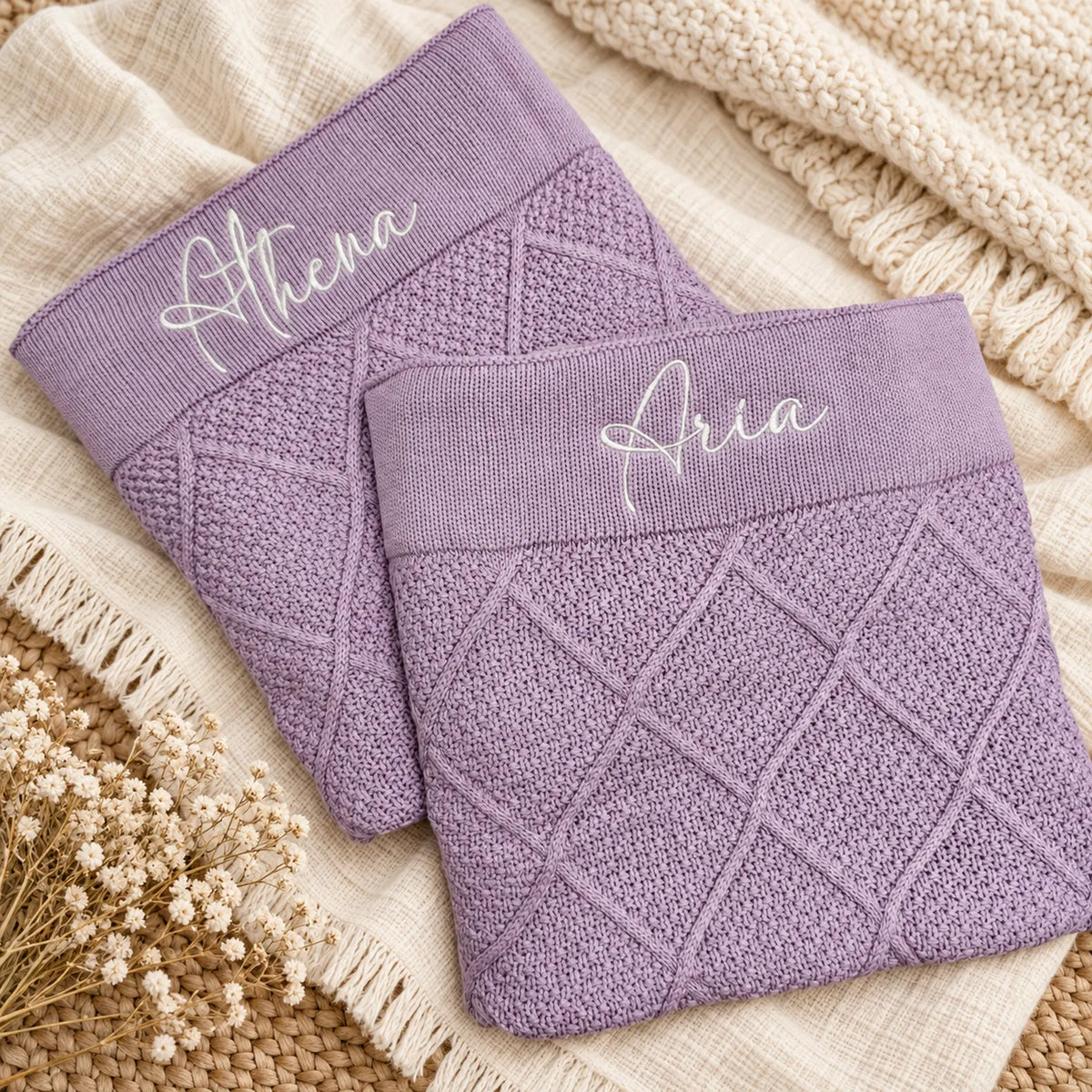 Two lavender knit blankets with diamond cable patterns and white embroidered names 'Athena' and 'Aria' on the cuffs, styled on a beige textured blanket with dried flowers.