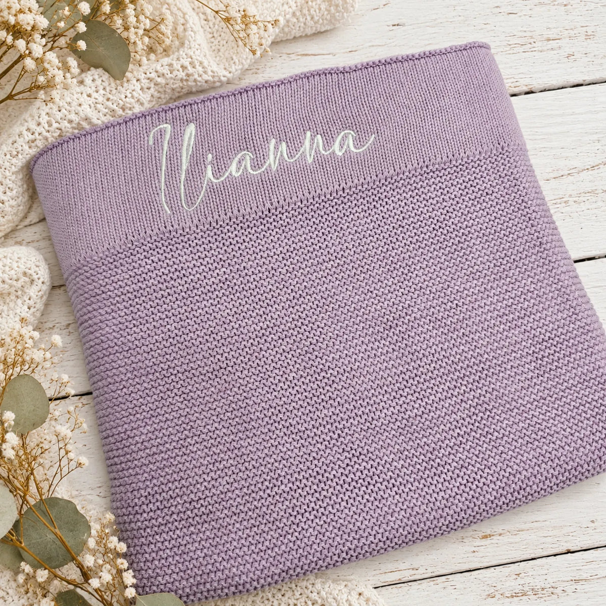 A lavender knit blanket with the name 'Iianna' embroidered in white script across the top, styled on a white wooden surface with dried flowers and a cream knit blanket.