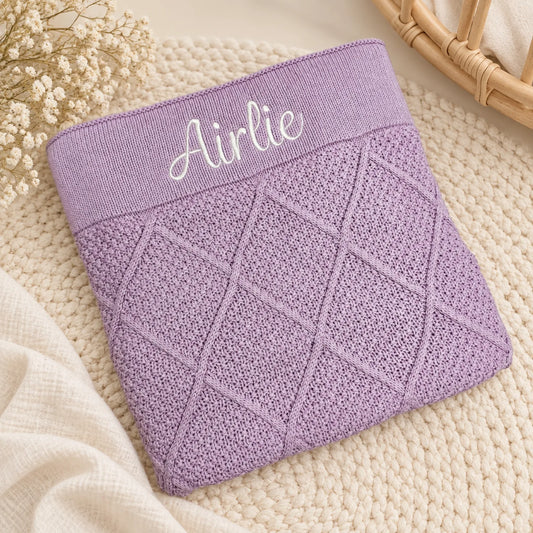 A folded lavender knit baby blanket or swaddle with a diamond cable-knit pattern and an elastic waistband embroidered with the name 'Airlie' in white script, styled on a cream knit blanket with dried flowers.