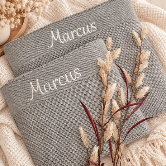 Two gray woven blankets or throw pillows with the name 'Marcus' embroidered in white script on the waistbands, styled on a cream knit blanket with dried grasses.